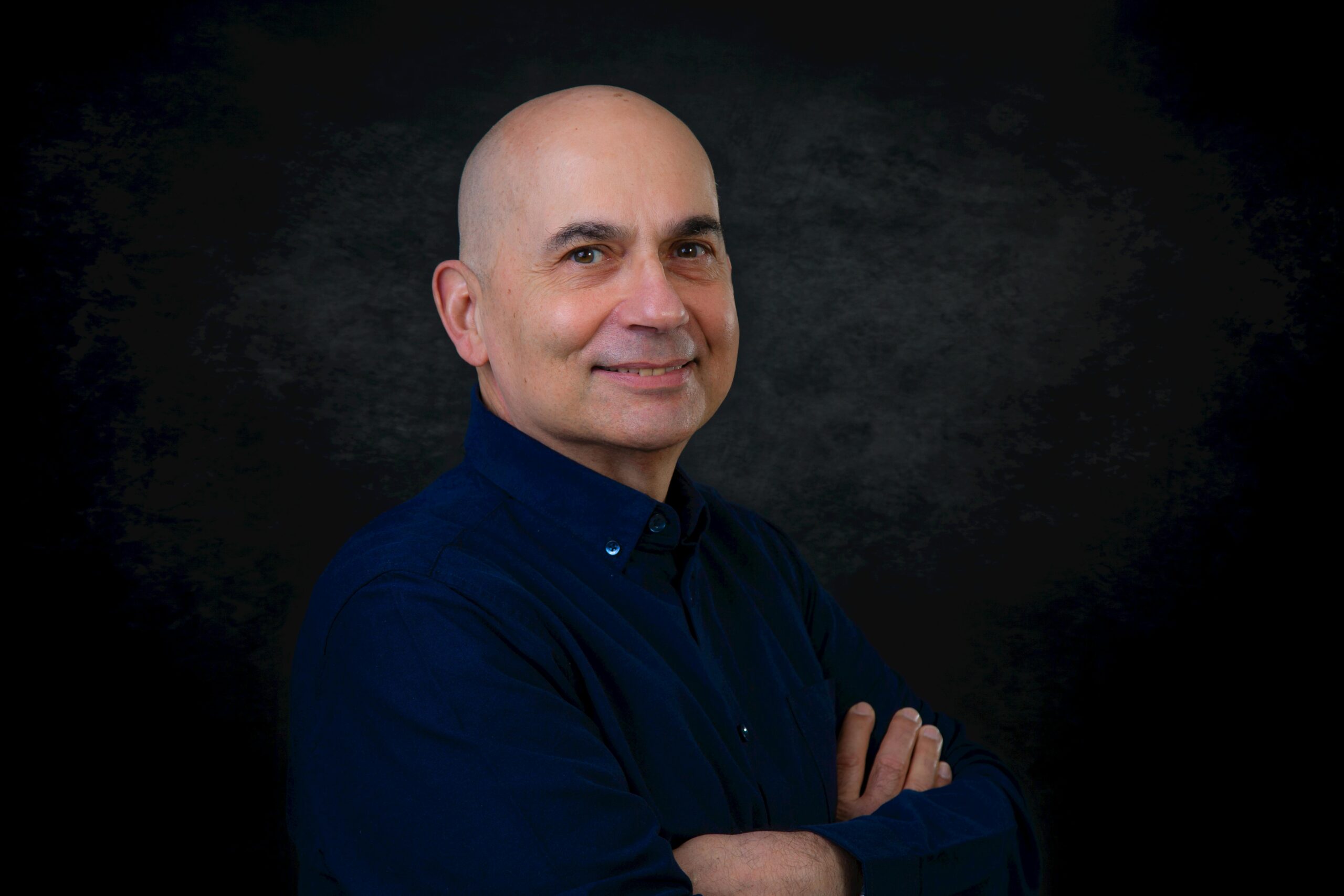 Bald man wearing a blue shirt, smiling confidently with crossed arms in a studio setting.