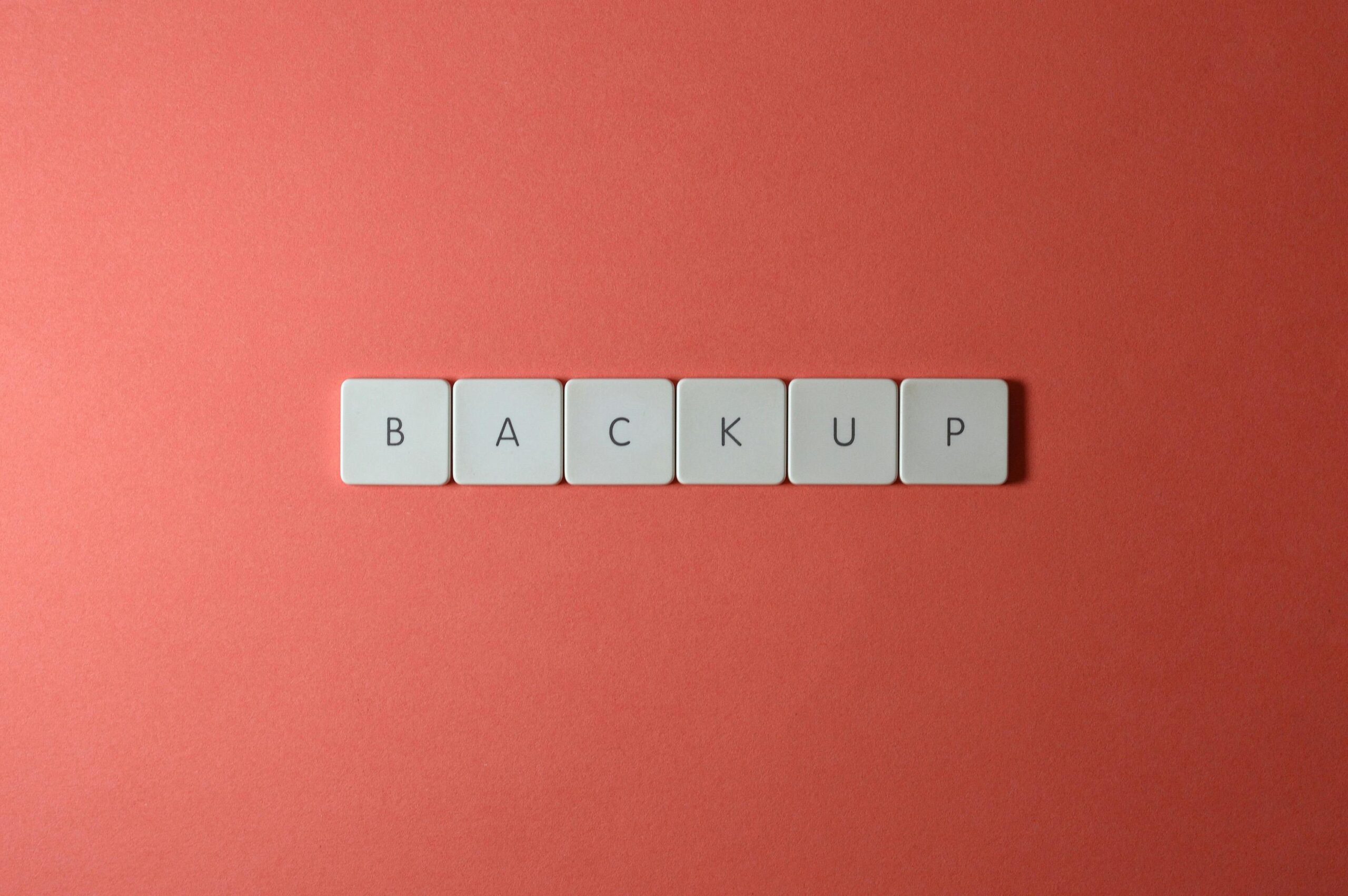 Close-up of keyboard keys spelling 'BACKUP' placed on a coral-colored surface.